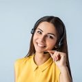 CUSTOMER Photo of beautiful young call center operator standing near gray background. Woman with headphones looking at camera and smiling